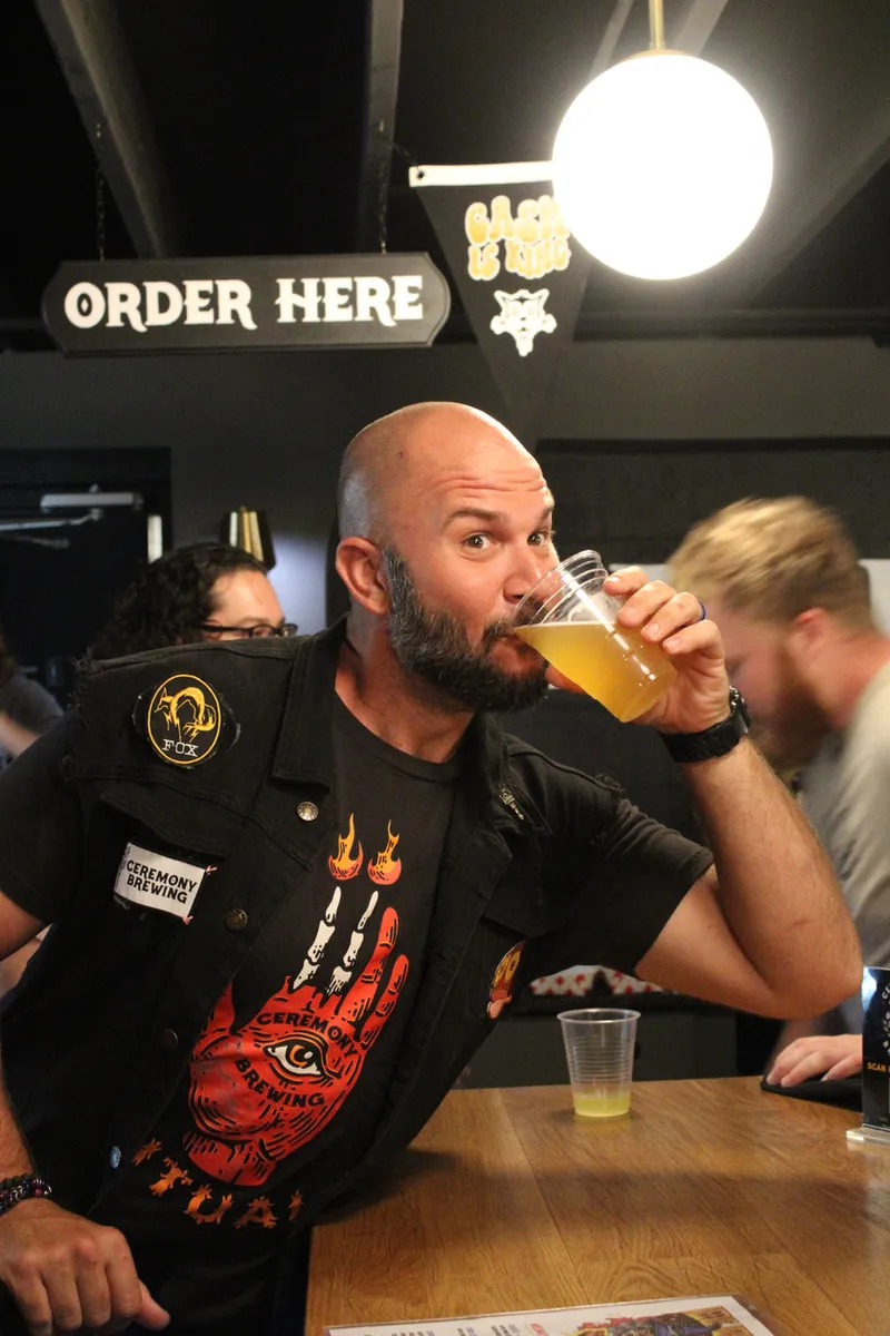 A Ceremony Brewing regular in a band vest enjoying a beer under the neon Order Here sign in the taproom